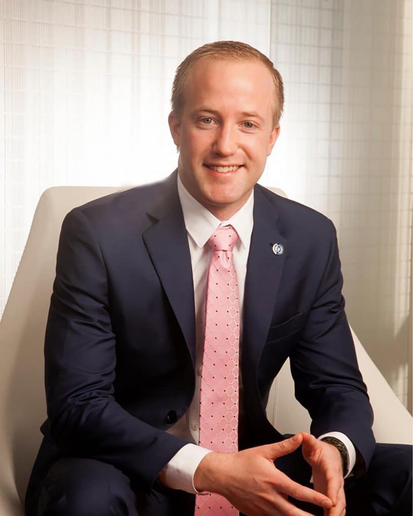 Man in a navy suit and pink tie seated in a white chair beside a bright window