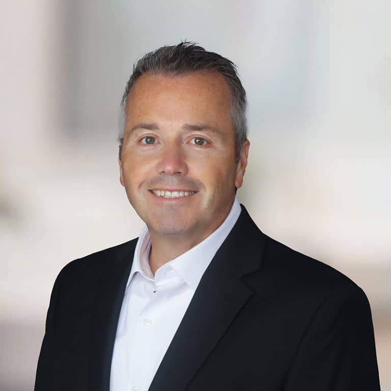 Man in a dark suit and open-collar shirt against a bright, softly blurred office
