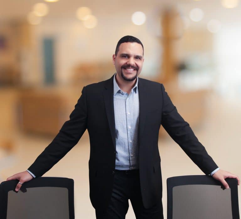 Man in a dark suit leaning on two office chairs, softly blurred office behind him