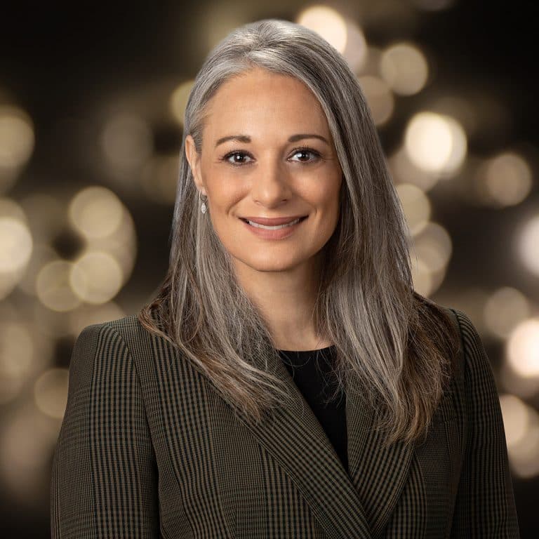 Woman with long silver hair in a tweed blazer, warm blurred lights behind her